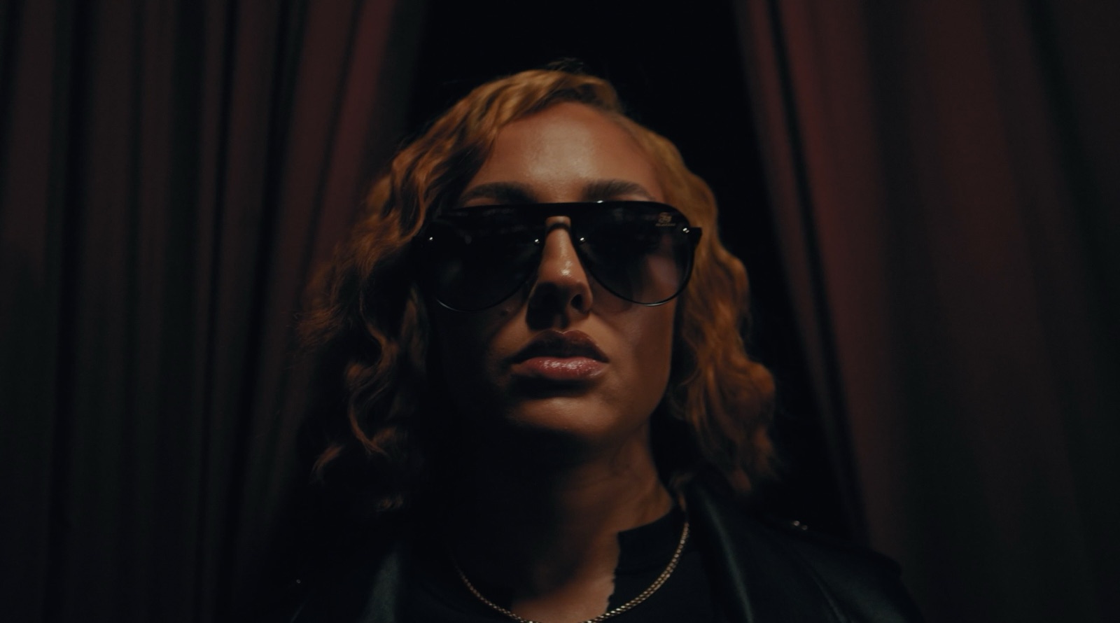 A woman in dark aviator sunglasses stands between red curtains, lit from above. Still from My Cherie Amour.