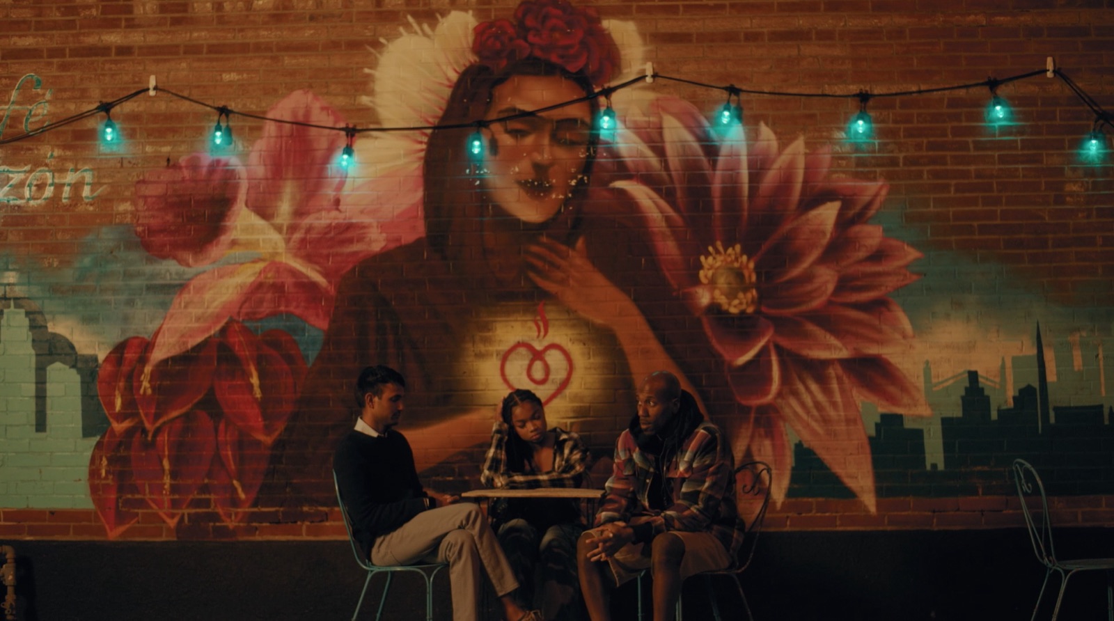 Three people sit at a cafe table beneath a Frida Kahlo mural strung with teal lights. Still from My Cherie Amour, courtesy of AGA Productions.